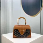 Front view of a rigid, structured handbag (Goyard Saïgon style) featuring a signature chevron/Y-pattern, wooden top handle, and corner reinforcements.