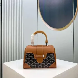 Front view of a rigid, structured handbag (Goyard Saïgon style) featuring a signature chevron/Y-pattern, wooden top handle, and corner reinforcements.