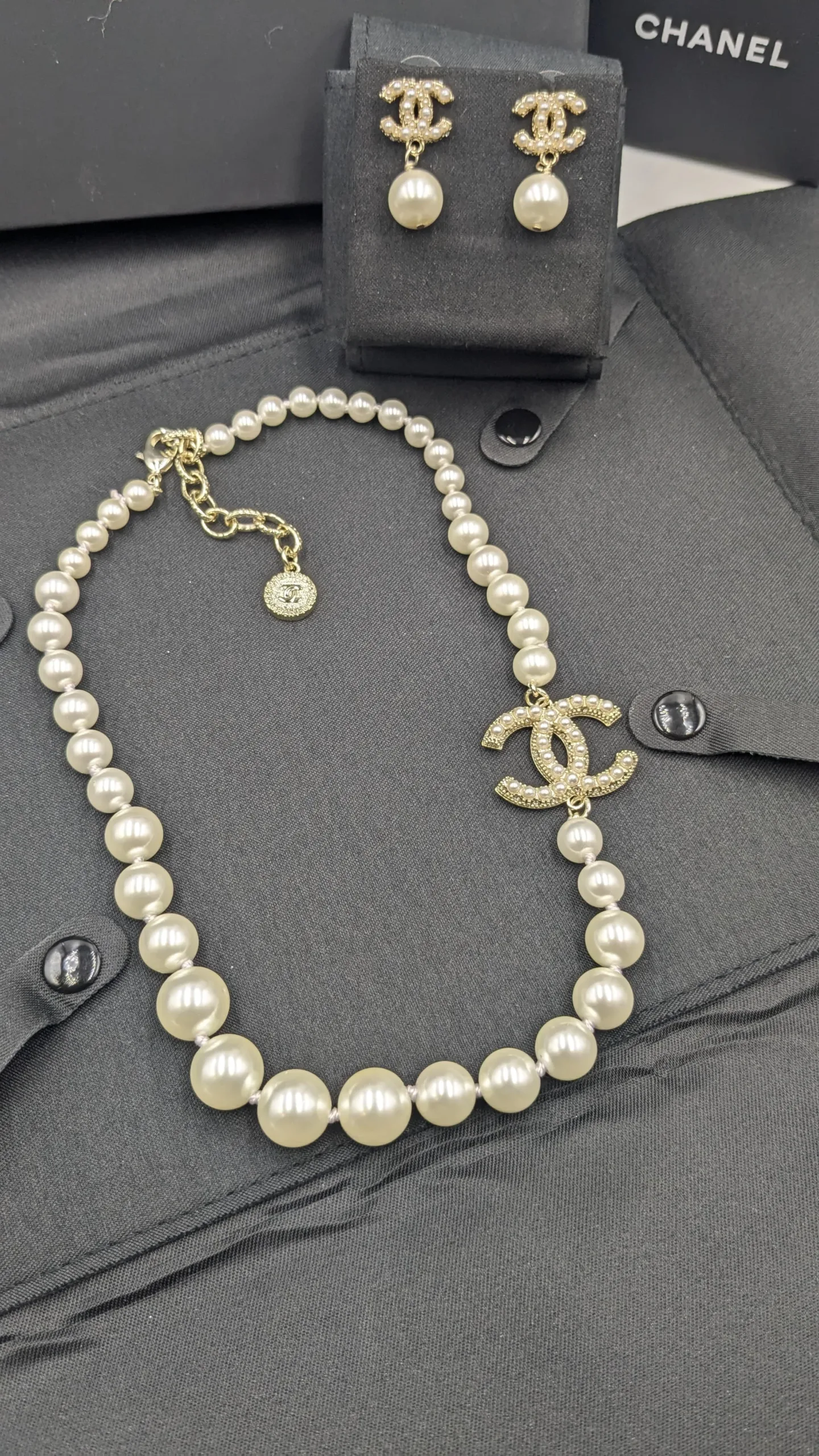 Chanel Inspired Pearl Necklace and CC Earrings Jewelry Set High-angle shot of a classic faux pearl necklace with a dazzling crystal-studded CC logo pendant, displayed with matching logo stud earrings on a black garment.