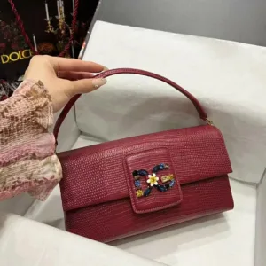 Red textured leather baguette shoulder bag featuring a multi-colored jeweled floral buckle detail, held by a hand.