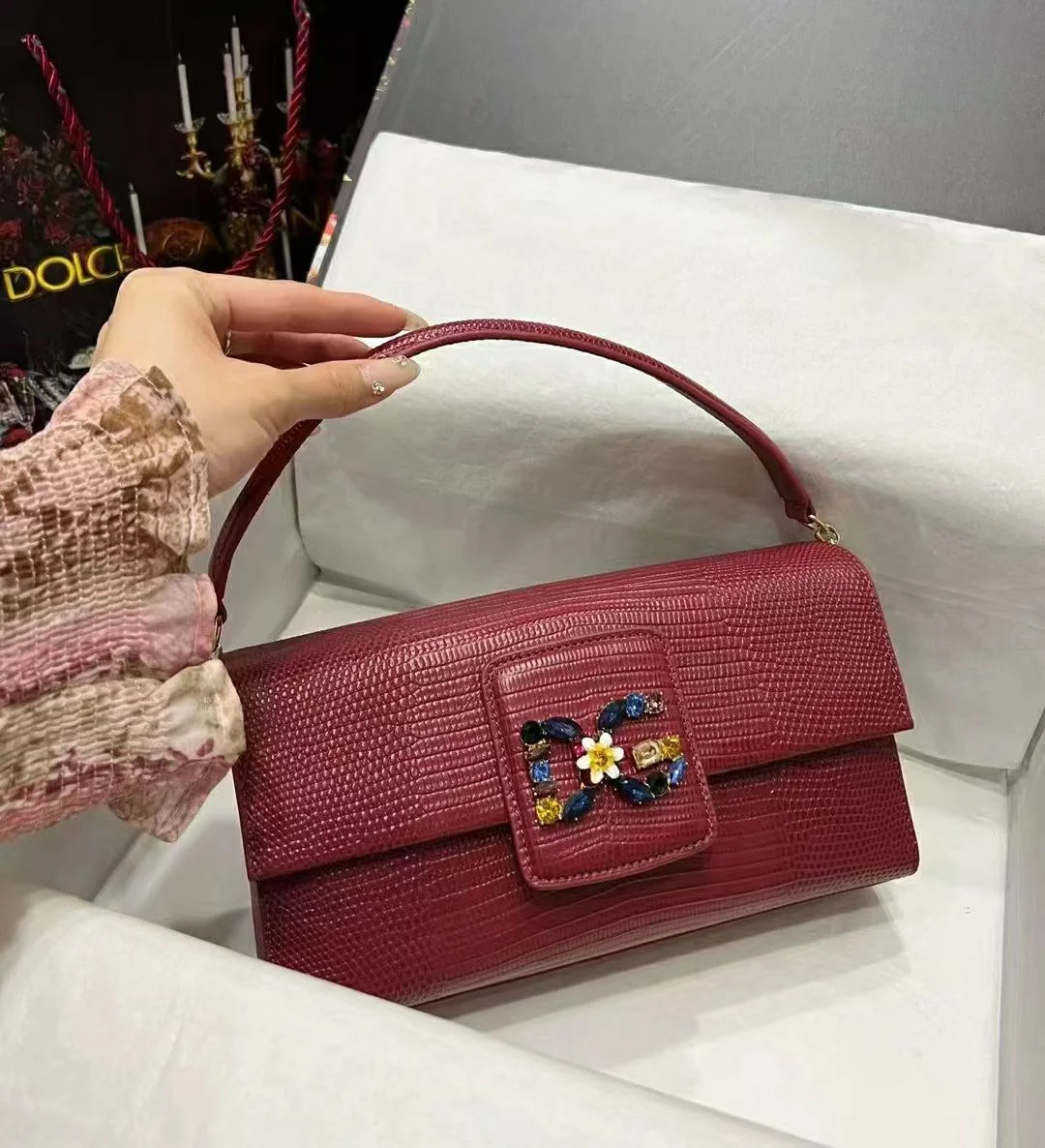 Red-Textured-Leather-Jeweled-Baguette-Bag Red textured leather baguette shoulder bag featuring a multi-colored jeweled floral buckle detail, held by a hand.