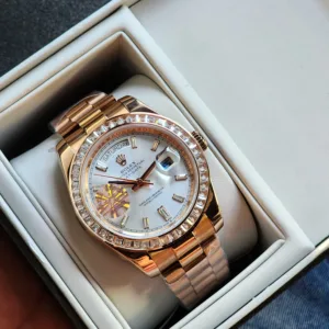 A rose gold tone Rolex Day-Date watch with a diamond bezel and a light-colored dial, resting in its presentation box.