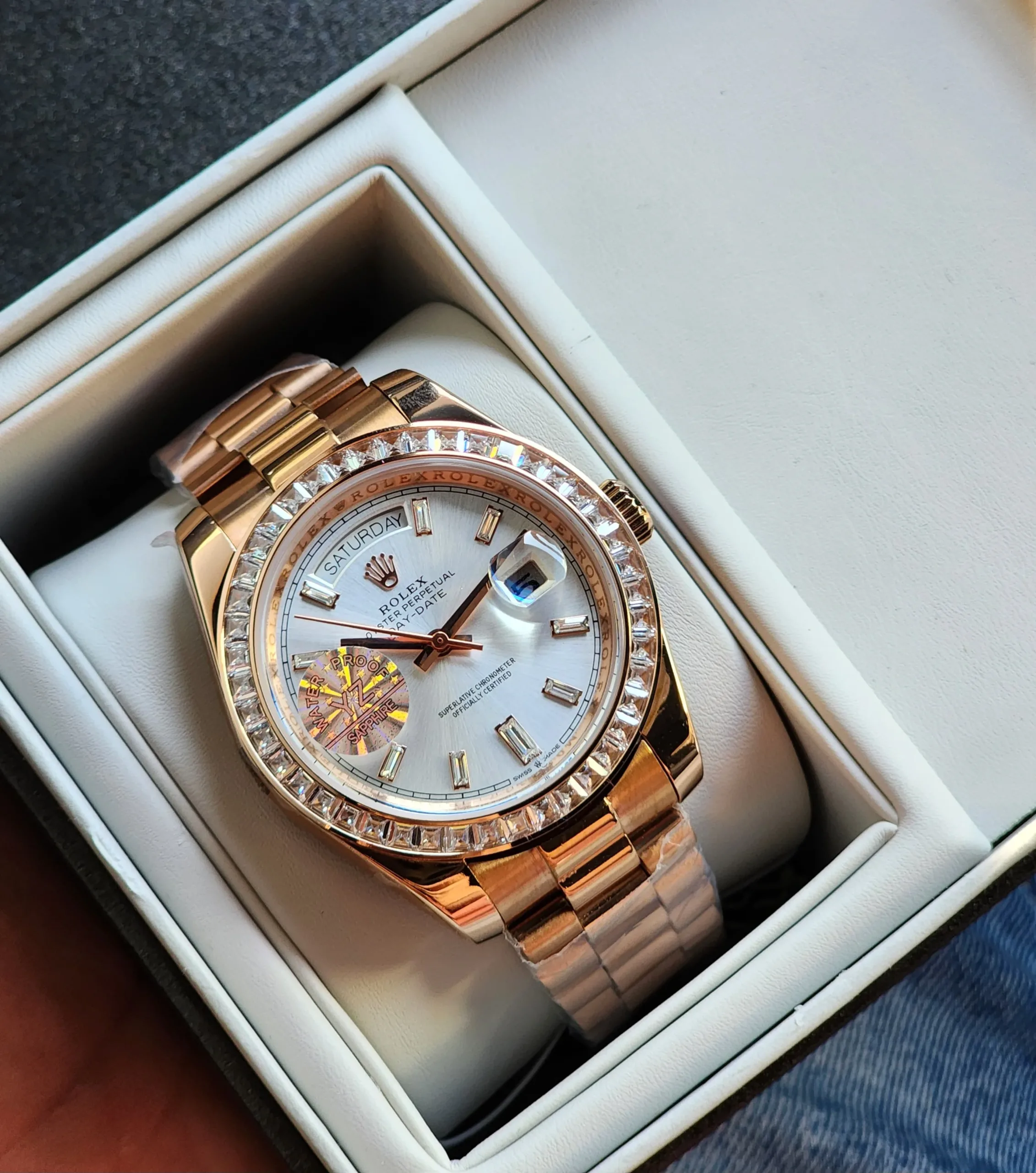 rolex-day-date-40mm-diamond-bezel-rose-gold A rose gold tone Rolex Day-Date watch with a diamond bezel and a light-colored dial, resting in its presentation box.