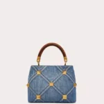 Front view of the Small Valentino Garavani Roman Stud Handle Bag in quilted denim with contrasting stitching and antique brass maxi studs.