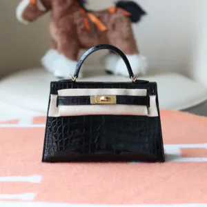 Close-up of shiny, structured mini crocodile handbag with gold-tone turn-lock hardware and top handle, resting in a white box.